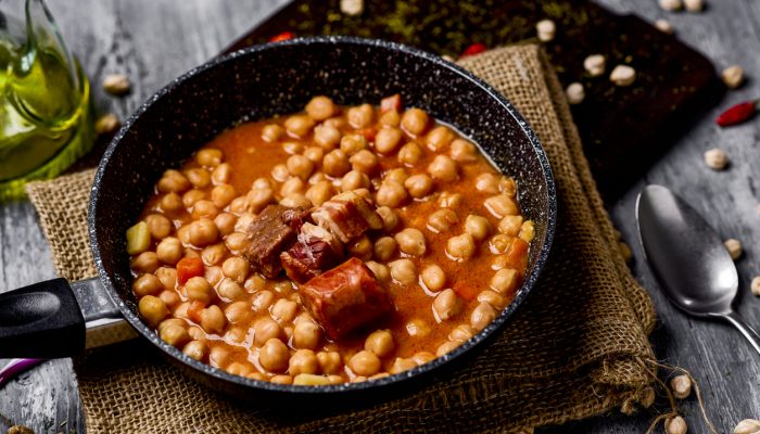 a stone frypan with garbanzos a la riojana, a spanish chickpeas stew, on a wooden chopping board placed on a rustic wooden table next to a cruet with olive oil